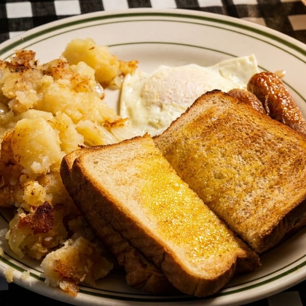 Breakfast plate with fried eggs, sausage links, home fries, and buttered toast on a checkered tablecloth.